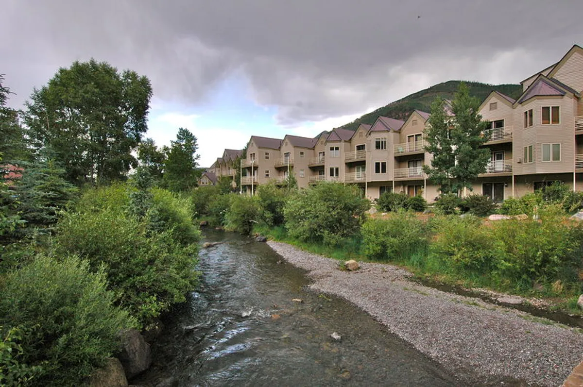 A tranquil view of the stream running alongside the buildings, surrounded by lush trees and well-maintained balconies. The scene suggests relaxation and easy access to nature, with residents walking along the stream and enjoying the outdoors from their balconies.