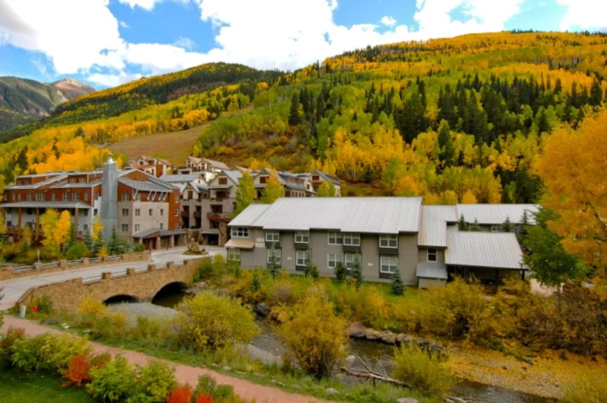 A vibrant autumn scene showcasing colorful foliage, a modern residential building, a meandering stream, and a picturesque bridge. The setting is tranquil, with nature and pathways inviting for exploration and relaxation by the water.