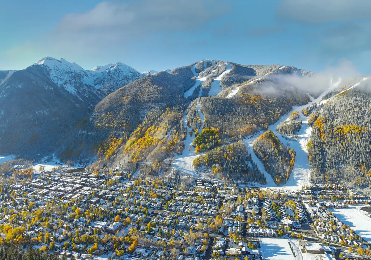 This image offers a sweeping view of snow-covered mountains, ski slopes, a vibrant mix of autumn colors, and the town of Telluride nestled below. It captures the proximity to both nature and the town, with visual cues linking outdoor adventure (skiing, snowboarding, hiking) to the lively local scene and festivals.