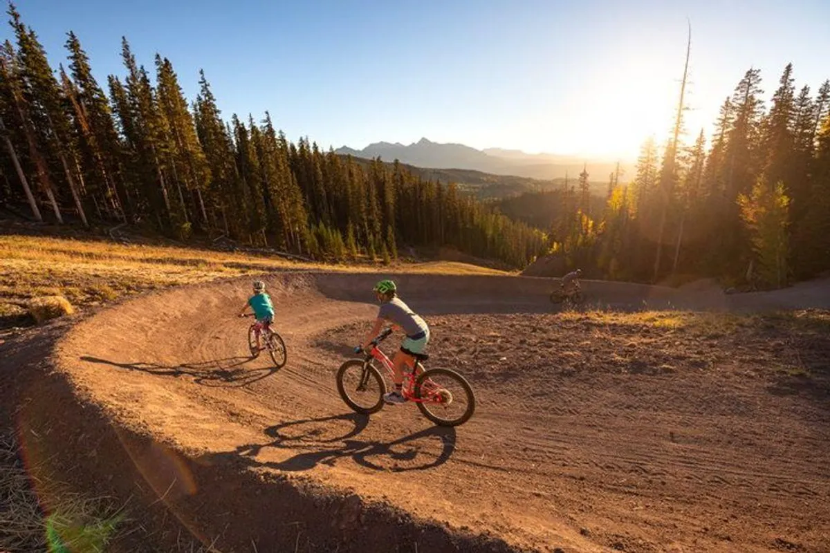 Adventurous mountain bikers navigate a winding trail at sunset through a forested landscape. The golden light, energetic movement, and natural setting exemplify outdoor excitement and the variety of adventure activities available beyond winter sports.