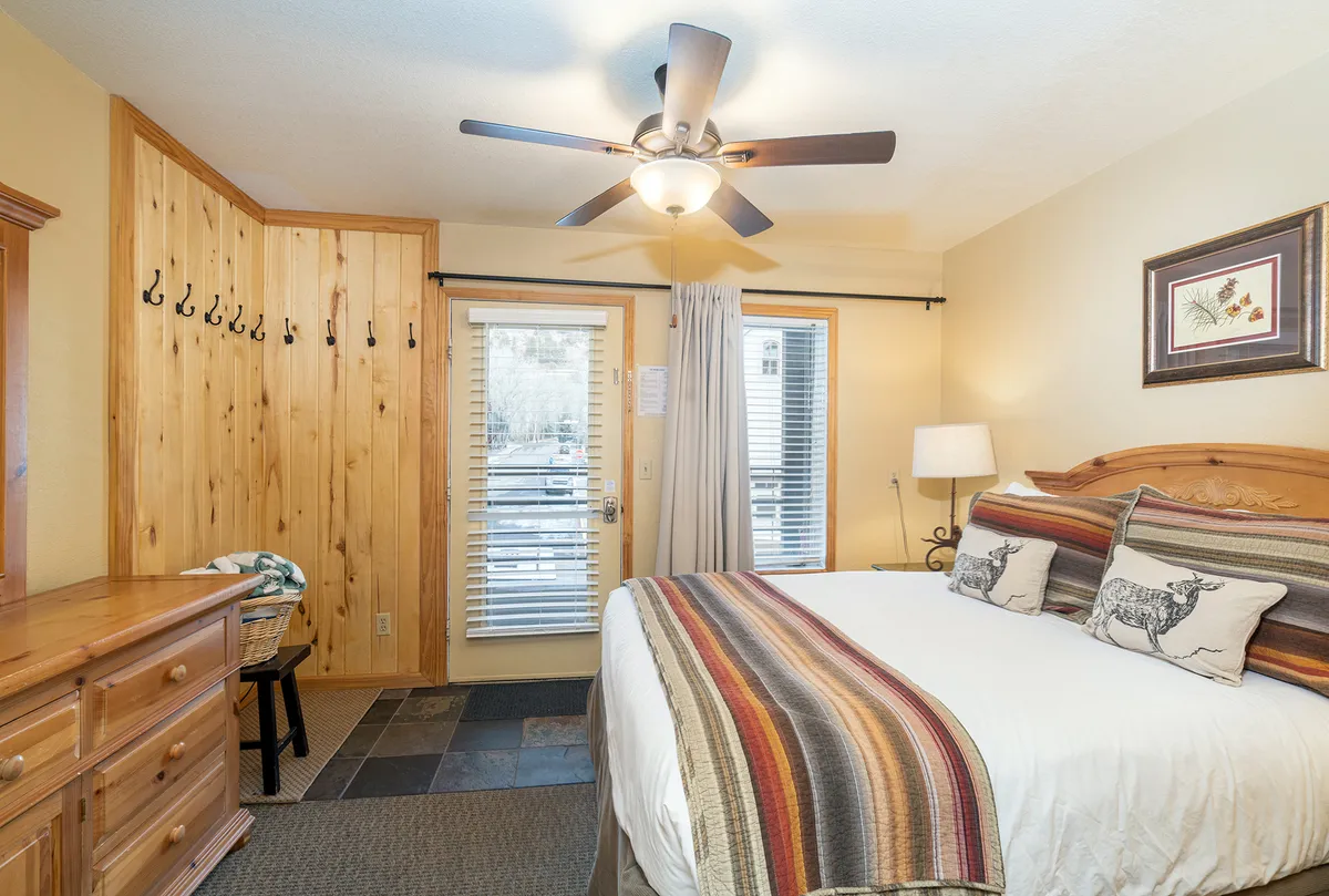 This photo features the queen bedroom with its colorful striped bedding, a wooden dresser, a door leading outside, and wall hooks for convenience. The space exudes warmth and natural light, offering an inviting, cozy retreat.